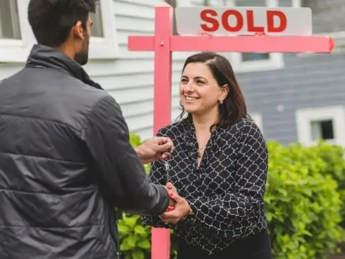 An estate agent handing house keys to a buyer in front of a sold sign